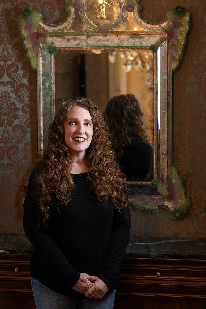 Person with curly brown hair, wearing a black top, stands in front of an ornate mirror and damask wallpaper.              