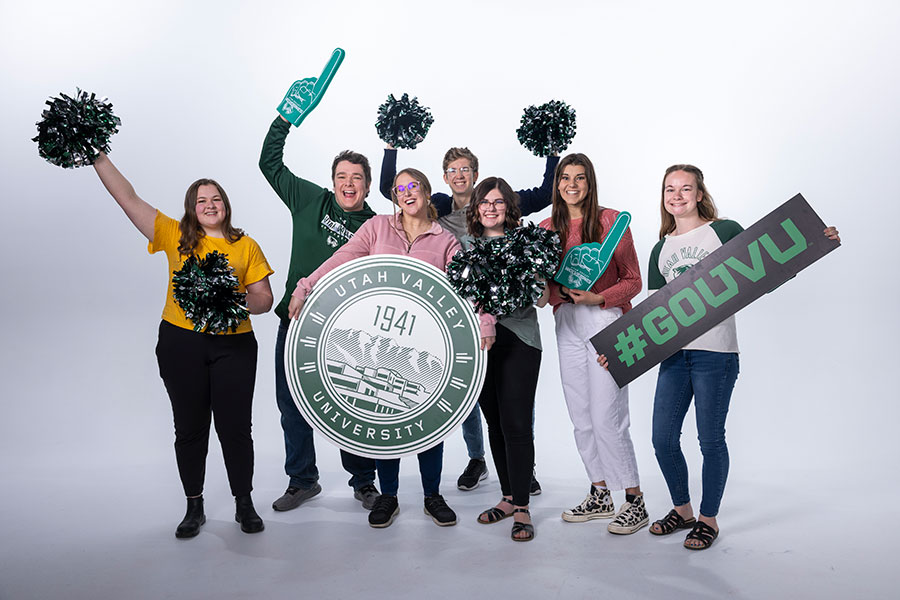 A group of seven people posing with pom-poms, foam fingers, and a university emblem.