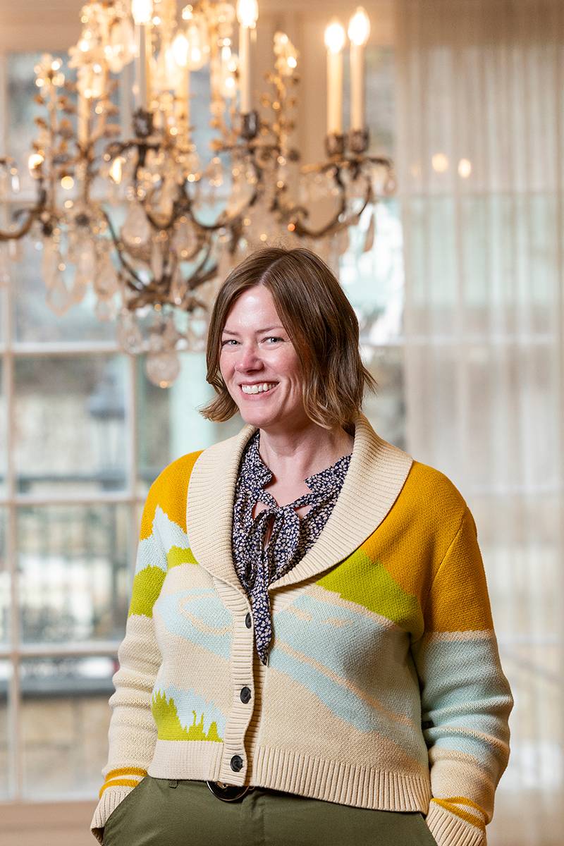 A woman wearing a colorful cardigan stands in front of a chandelier.              