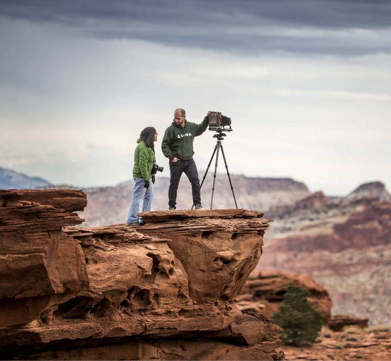 Capitol Reef Two people on a red rock cliff, one adjusting a camera on a tripod