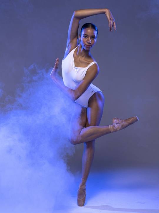 A ballet dancer in a white leotard performing on point surrounded by blue and purple mist