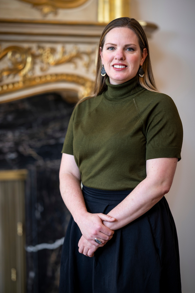 Person standing in front of an ornate fireplace with gold detailing.              