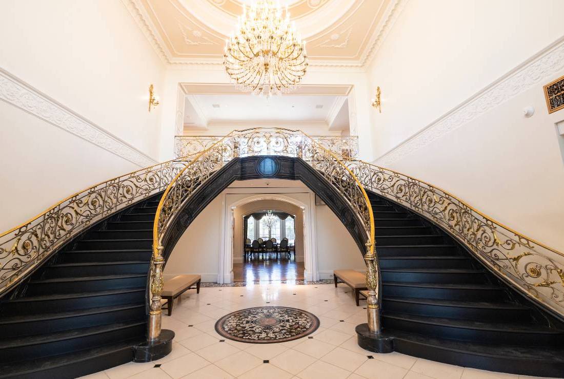 Grand staircase with dark wood, gold railings, white tile flooring, and a large chandelier above.              