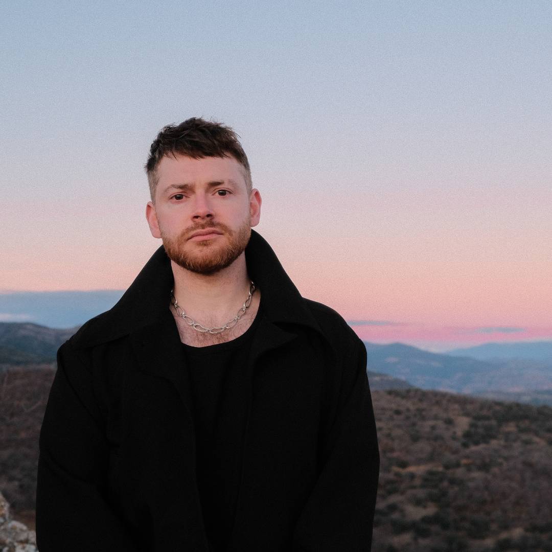 A man with short hair and a beard wearing a black coat and chain necklace stands against a pastel sunset sky and rolling hills.              