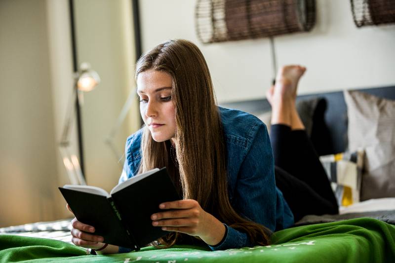 female student laying on her bed, reading a book