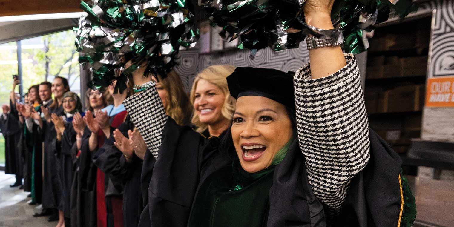 President Tuminez waves green and silver pom-poms and cheers while standing with faculty members and Board of Trustee members during UVU’s 2024 commencement ceremony processional. 