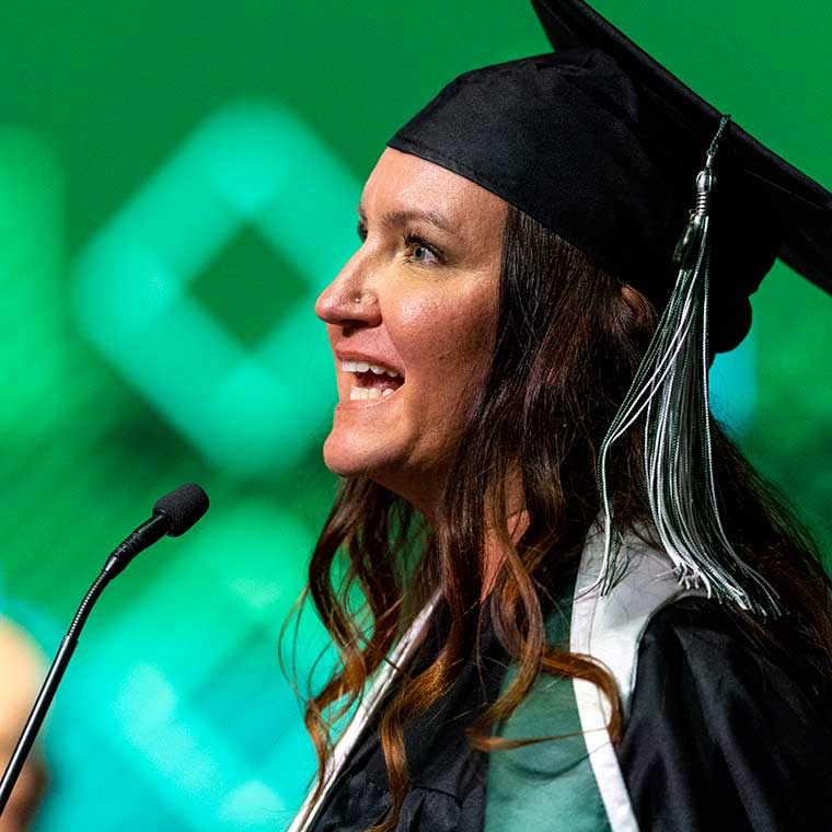 A student with brown hair speaks at a podium while wearing graduation regalia. 