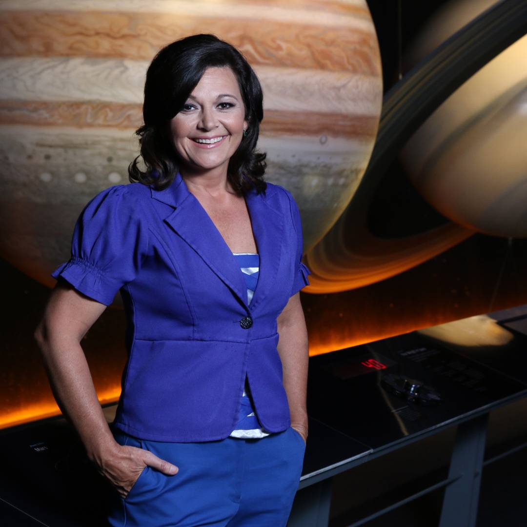 A woman in a purple blazer stands in front of large images of Jupiter and Saturn.