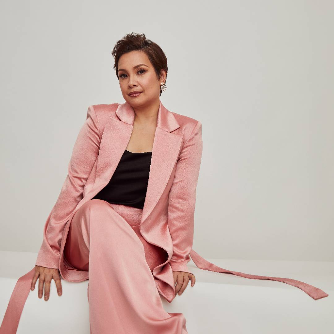 Person seated in a pink satin suit with a black top in a minimalist setting.              