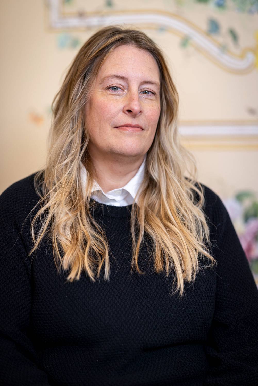 A person with long blonde hair in a black sweater and white shirt, set against a blurred floral background.              