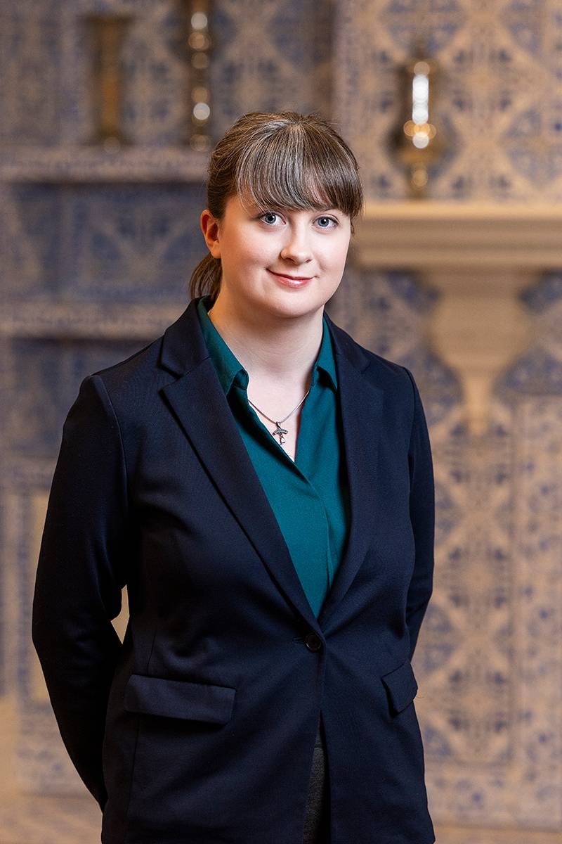 Person wearing a dark blazer and teal shirt standing in front of blue and white patterned tiles.              