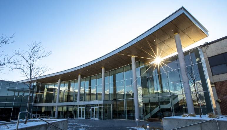 A modern building with a curved glass facade, tall columns, and sun streaming through one section.