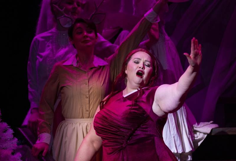 Opera Performance A women in a red dress sings on stage with other performers in the backgrounod under purple lighting