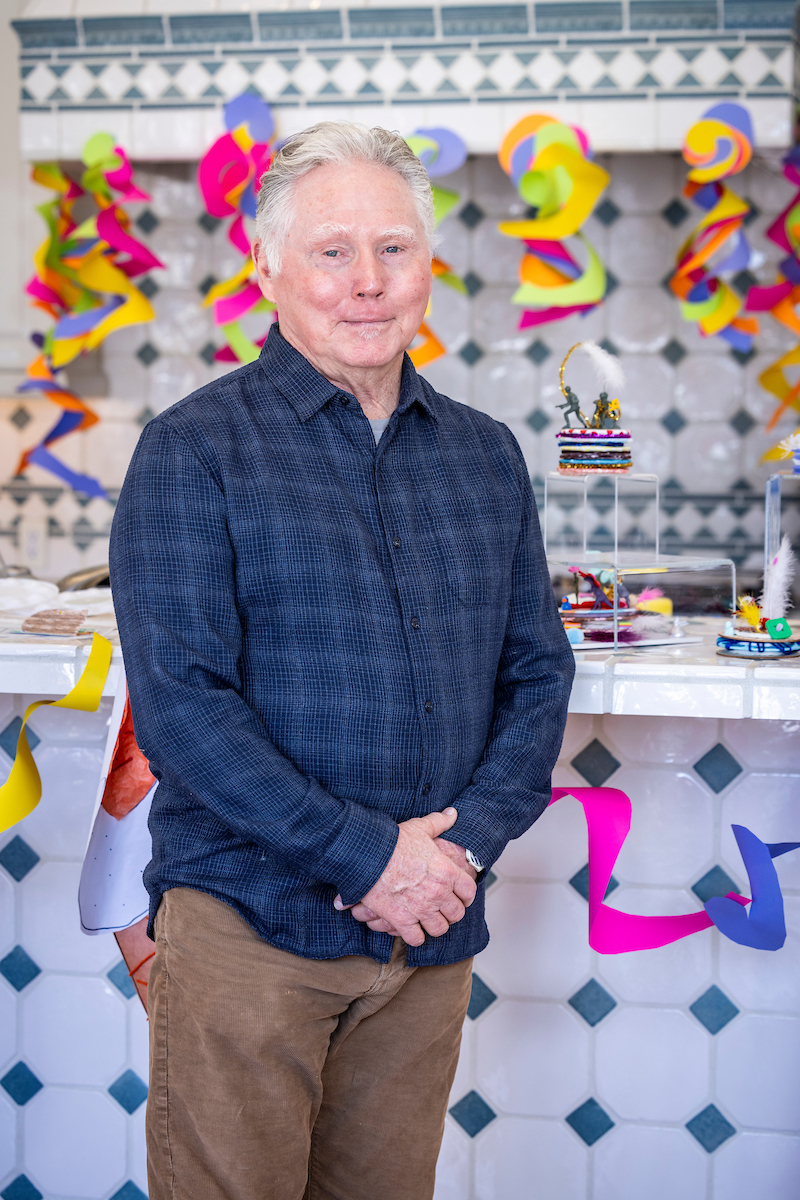 An older man in a blue checkered shirt stands in front of a colorful paper decoration background.              