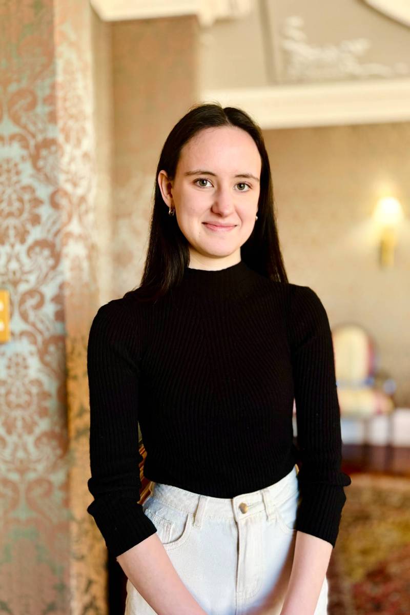 A person with long dark hair wearing a black top and light high-waisted pants stands in a room with ornate wallpaper.              