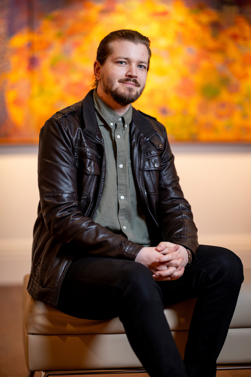 Person seated on a bench wearing a leather jacket, with abstract, colorful artwork in the background.              
