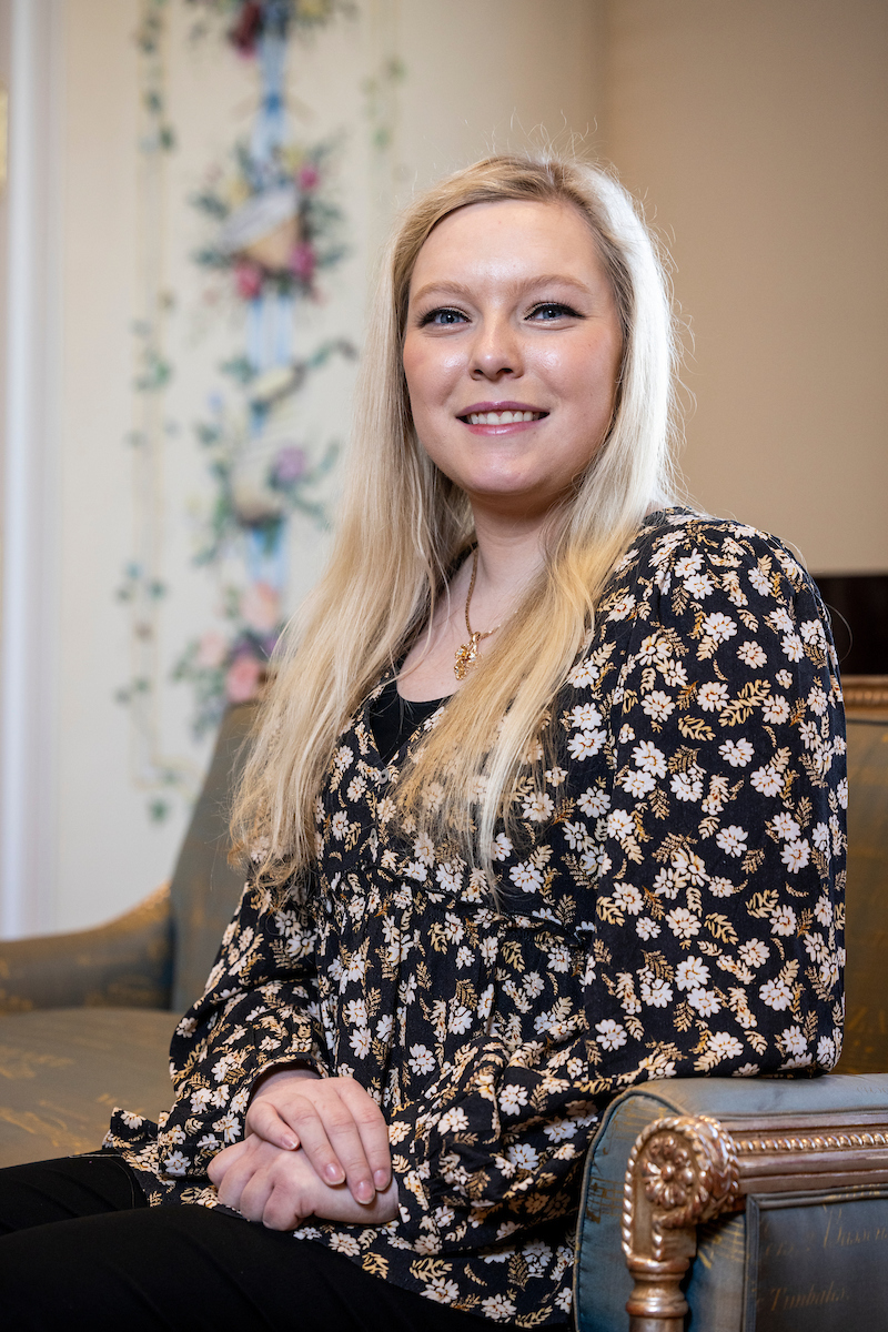 A woman with blonde hair in a floral dress sitting on a decorative chair.              
