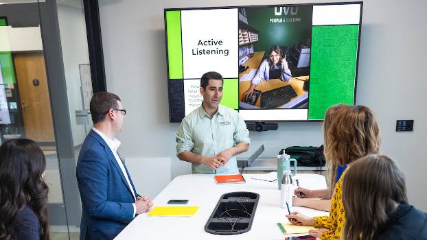Five people around a conference table. One person is at the front of the group presenting.