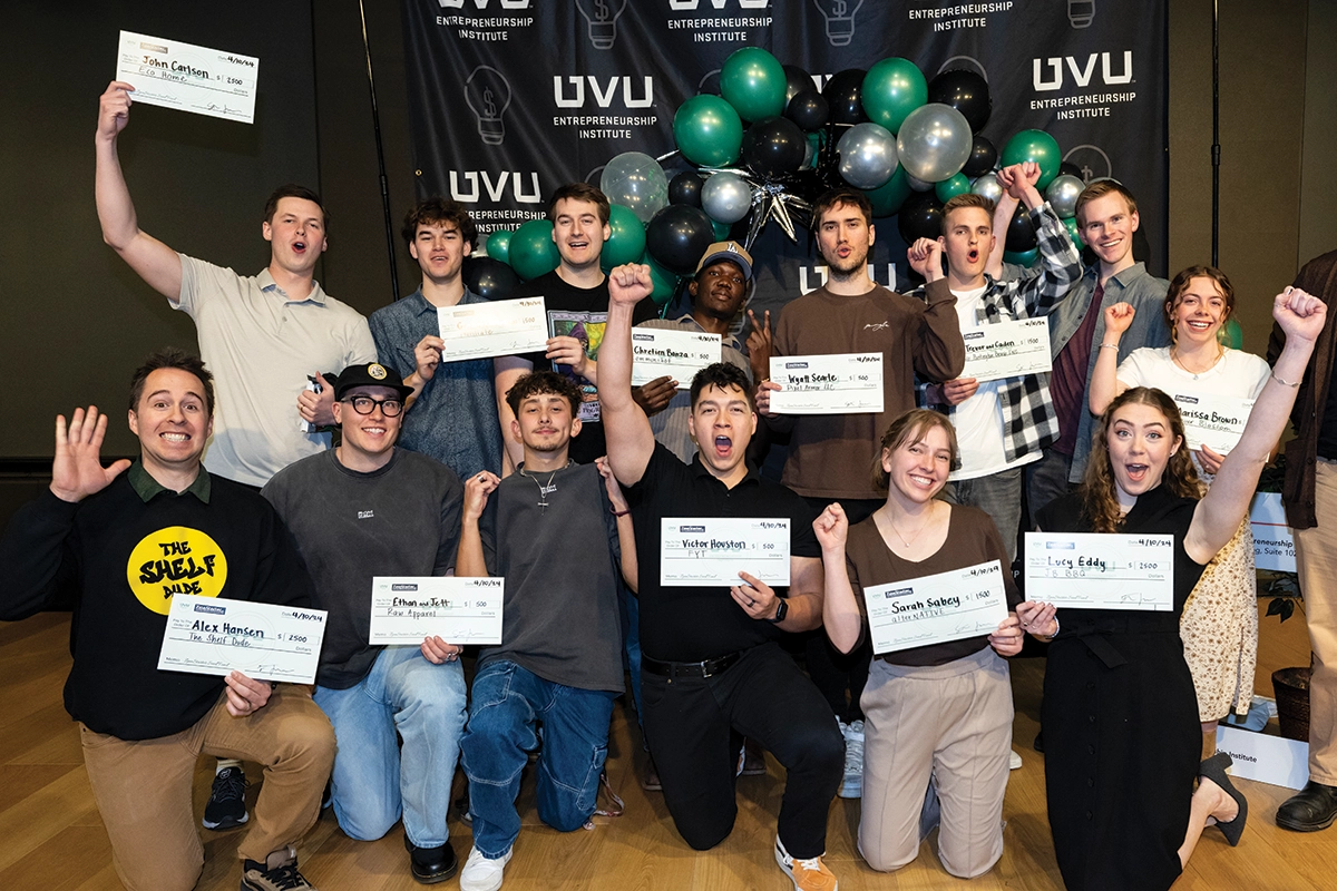 UVU Entrepreneurship Institute students pose and cheer against a black UVU backdrop while holding seed funding checks they won in a competition. 
