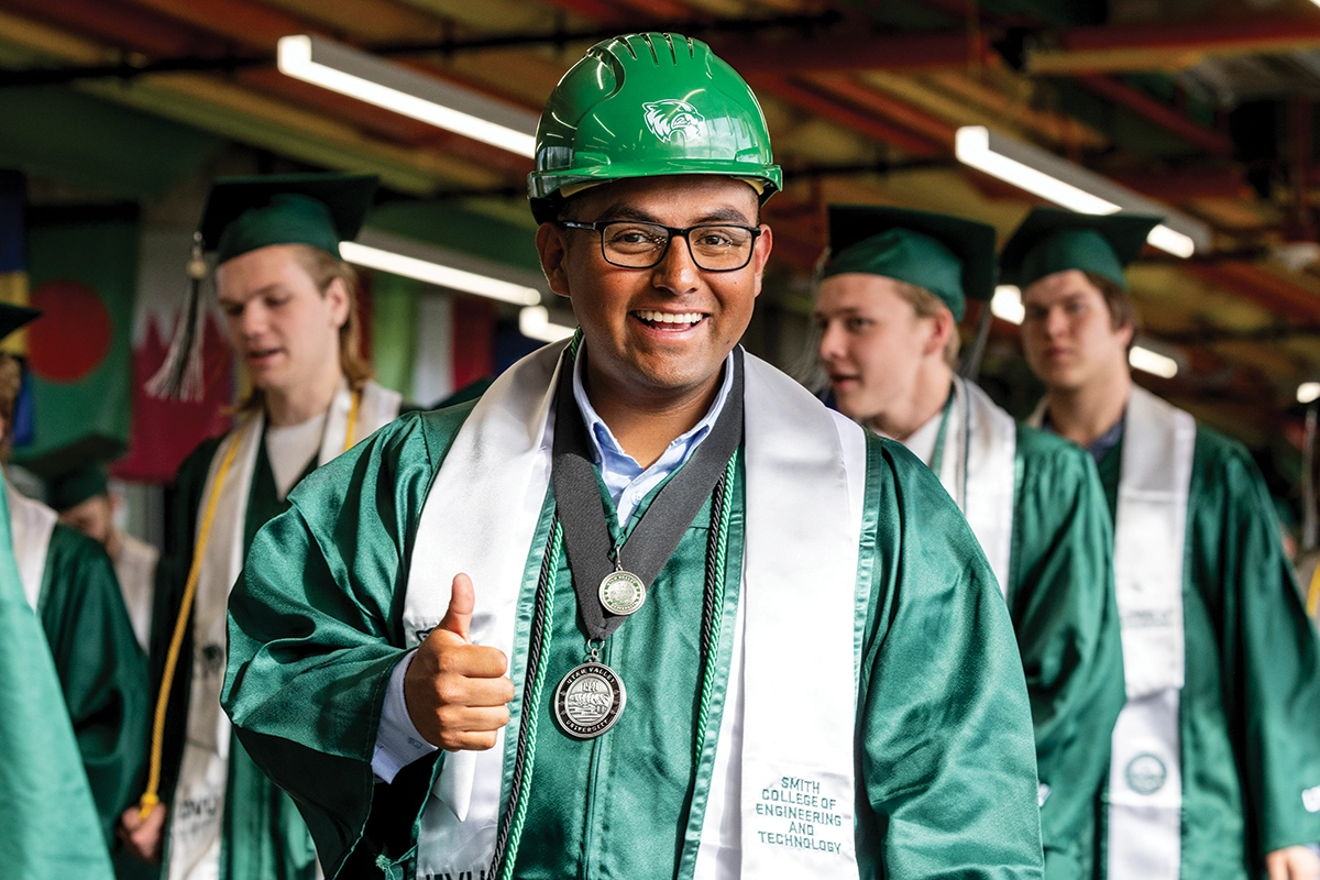A graduating student from UVU’s Smith College of Engineering and Technology, wearing a green construction hard hat, green regalia, and two medals, smiles at the camera while giving the thumbs up. 