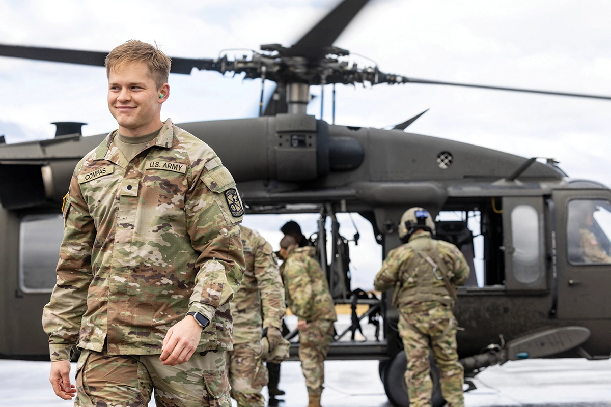 Four military-affiliated UVU students, wearing camouflage Army gear, walk near a black helicopter.