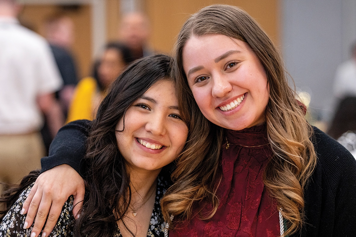 Two UVU students, one in a black floral blouse and the other in a red blouse, smile at the camera, one with their arm over the other’s shoulders. 