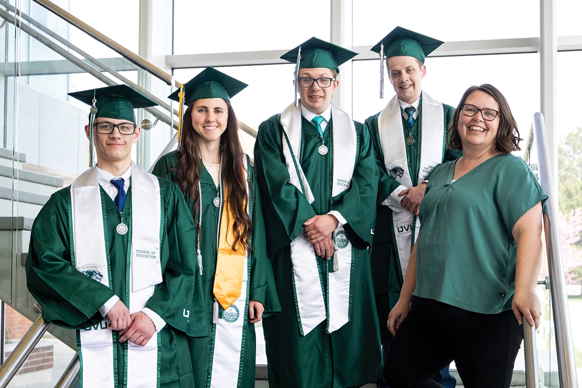 Graduating students from UVU’s Wolverines Elevated program, wearing green regalia, pose on UVU campus stairs with a faculty member from the program. 