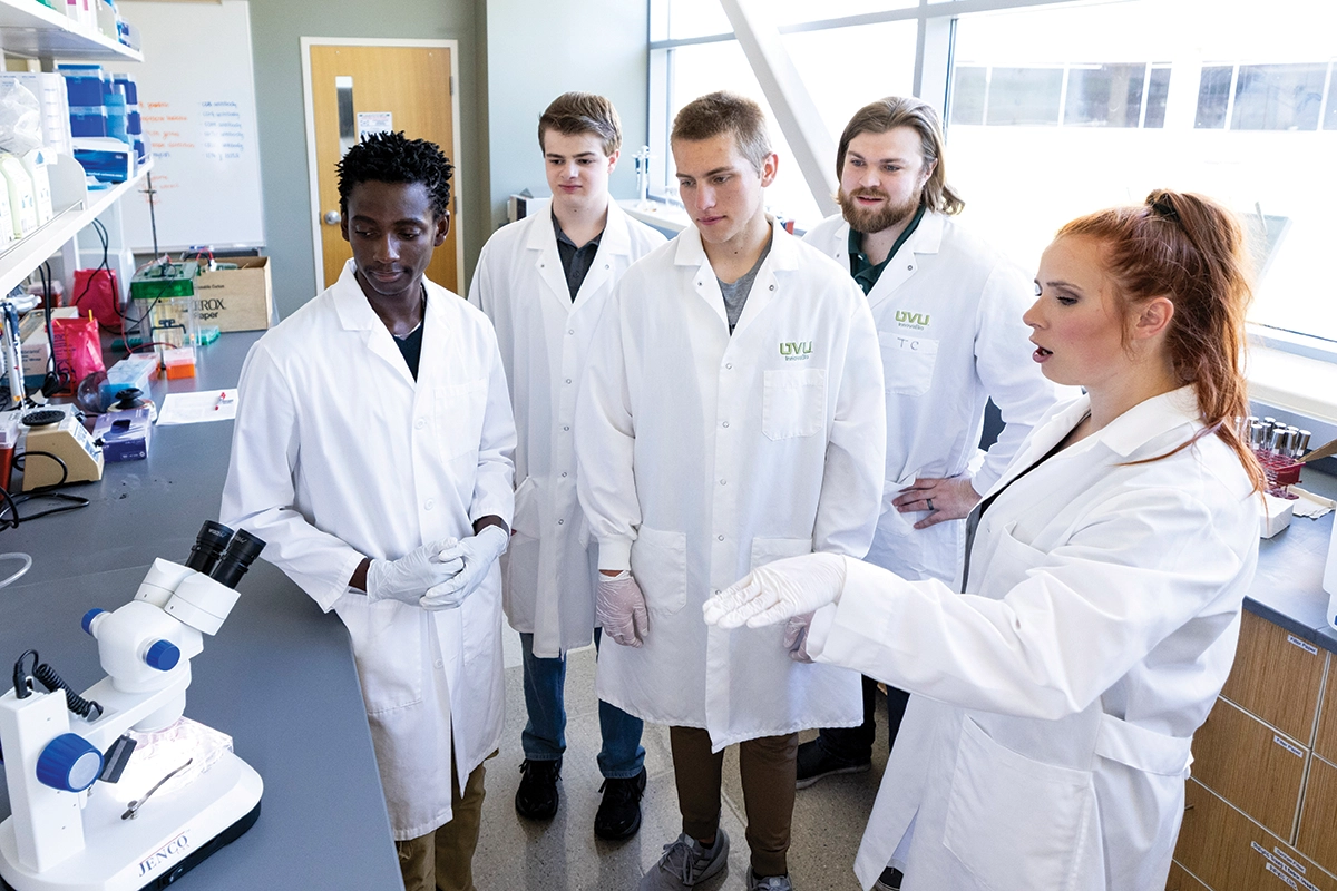 Five UVU students wearing white lab coats stand and talk in a laboratory. 