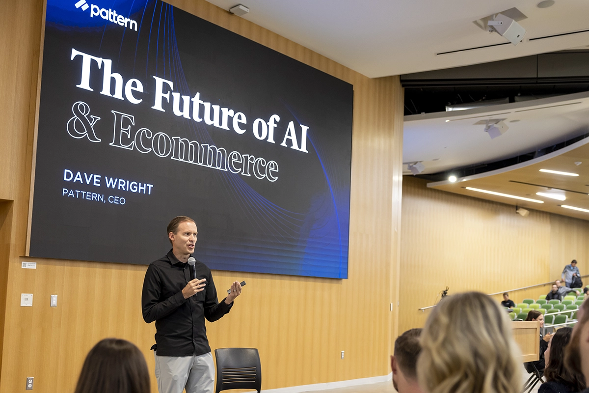 A person wearing a black shirt speaks into a microphone on a stage against a screen reading 'The Future of AI and Ecommerce.'