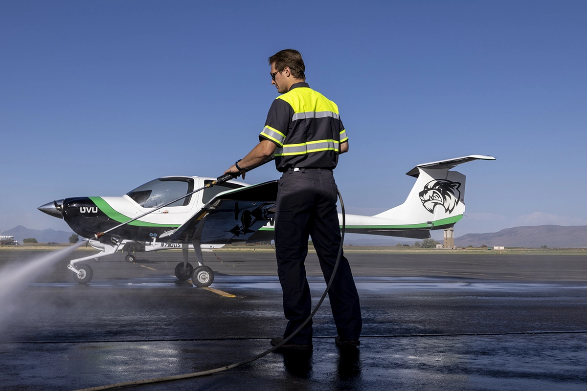 A student sprays a hose while standing near an airplane with the UVU Wolverine logo. 