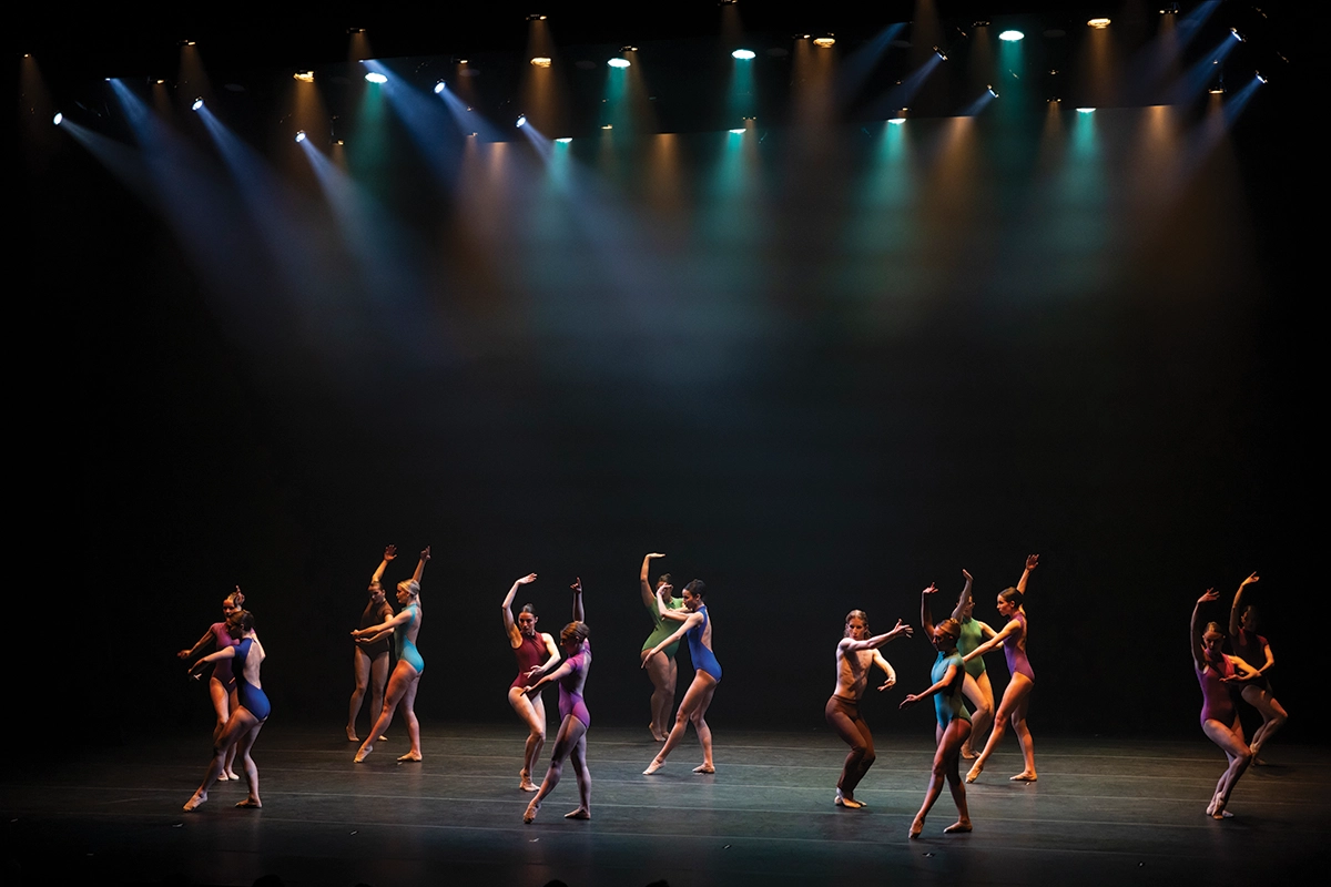Various UVU dance students wearing colorful leotards perform on a dark stage.