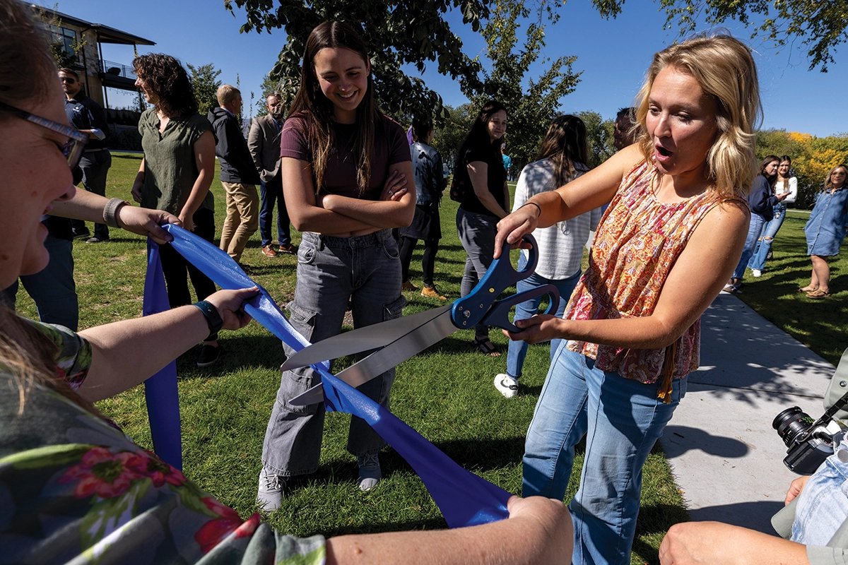 A person wearing a floral top and jeans cuts a blue ribbon with giant scissors at a community event promoting a new educational plant walk in Vineyard, Utah. 
