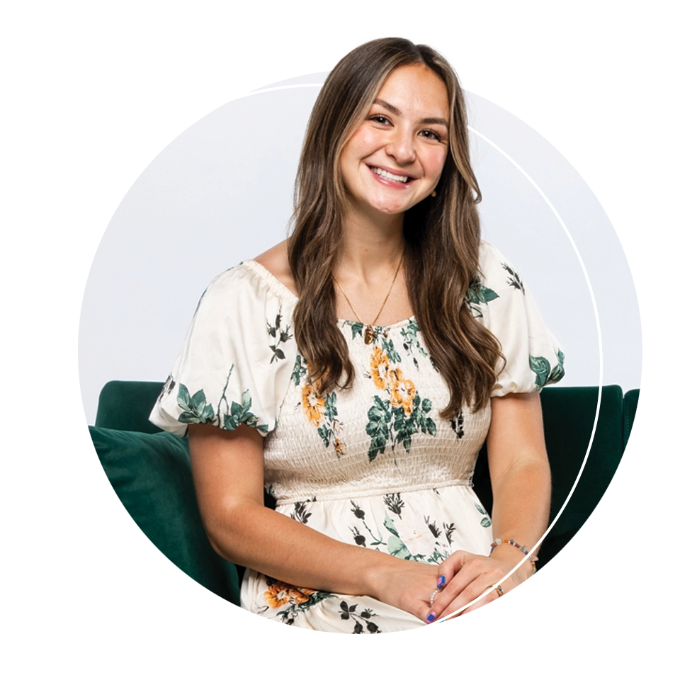 UVU alumna Maysen Rollo Minor, wearing a white floral dress, smiles while sitting on a dark green sofa. 