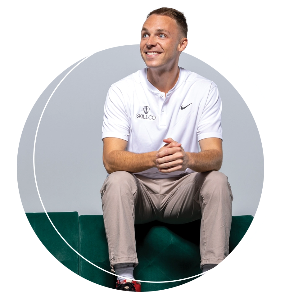 UVU alumnus Steve Lockhart, wearing a white shirt bearing the SkillCo logo, smiles while sitting on a green sofa. 